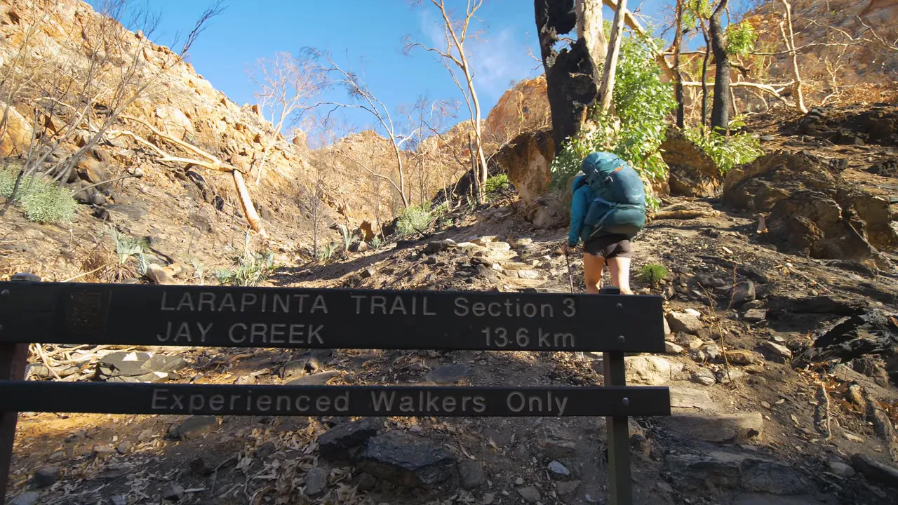 Larapinta Trail Section 3 Jay Creek sign with hiker climbing rocky trail; sign warns experienced walkers only